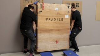 Filip Antonijevic und Manuel Suhr transportieren die große Klimakiste mit der Aufschrift „Fragile“ (zerbrechlich), die das Bild enthält.