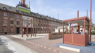 Vor dem Torhaus weisen ausgediente Schilder und Überwachungskameras auf die Geschichte des Ortes hin. Das Wachhäuschen wird gerade gestrichen.