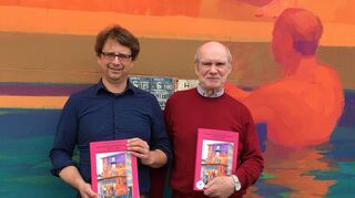 Philip Gerber (l.) und Wolf Preißner mit der Chronik zum Geburtstag des Drogenvereins. Im Hintergrund ist ein Ausschnitt des großen Wandbildes zu sehen, das der französische Künstler Zoer im vergangenen Oktober an das Gebäude in K 3 gesprüht hat und das eine Urlaubsszene an der Côte d’ Azur zeigt.