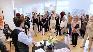 Prince-House-Gallery-Chef Johann Schulz-Sobez (am Mikrofon) begrüßt den Künstler Manfred Fuchs (rechte Bildhälfte vor der roten Skulptur) und die weiteren Gäste zur Vernissage der Fuchs-Benefizschau „Licht – Farbe – Erlebnisse“.