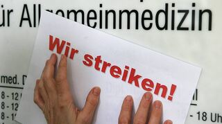 Am Eingang einer Arztpraxis befestigt eine Mitarbeiterin einen Zettel mit der Aufschrift „Wir streiken!“