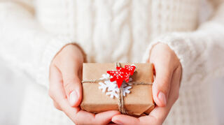 Woman in knitted sweater holding a present. Gift is packed in craft paper with white felt snowflake and red fir tree. DIY way to pack Christmas presents.