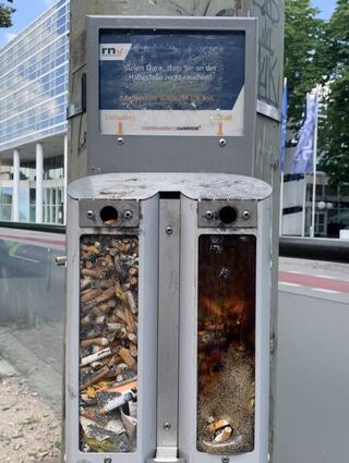 Volle Aschenbecher an einer Haltestelle. Das Plexiglas ist rechts geschmolzen.