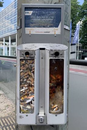 Volle Aschenbecher an einer Haltestelle. Das Plexiglas ist rechts geschmolzen.