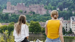 Zwei junge Frauen sitzen mit Blick auf das Heidelberger Schloss an der Alten Brücke.