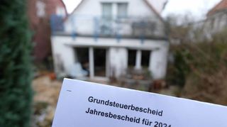 Ein Grundsteuerbescheid wird vor einem Privatgrundstück hochgehalten. Ab nächstes Jahr tritt die Grundsteuerreform in Deutschland in Kraft.