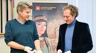 Lars Börner (l), Kurator, und Alexander Schubert, Museumsdirektor, zeigen ein ägyptisches Salbgefäss aus dem 1. Jahrhundert vor Christus vor einem Plakat zur geplanten Ausstellung zu Caesar und Kleopatra.