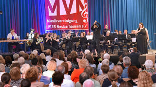 Krimi-Konzert der Musikvereinigung Neckarhausen: Dirigent Carlos Trujillo Mendez in James-Bond-Pose, rechts Sängerin Sophie Speicher.