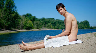 Mannheims Stadtrat Julien Ferrat sitzt mit dem Amtsblatt der Stadt Mannheim am Ufer der Friesenheimer Insel. Ferrat plant ein Trainingscamp am örtlichen FKK-Strand für einem FKK-Swinger-Urlaub in Frankreich.