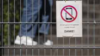 Ein Schild mit einem durchgestrichenen Smartphone und der Aufschrift «Auf dem ganzen Schulgelände - zu jeder Uhrzeit! DANKE!» hängt am Zugangstor zum Schulhof eines Gymnasiums und weist auf ein generelles Handyverbot hin.