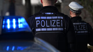 Das Logo der Polizei auf der Dienstkleidung von Polizisten, die vor einem Blaulicht stehen (gestellte Szene).