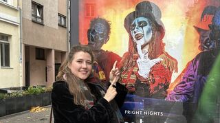 Lena Ritthaler vor dem Plakat, auf dem sie in der Rolle zu sehen ist, die sie seit 2019 zu den "Halloween Fright Nights" im Plopsaland Deutschland verkörpert.