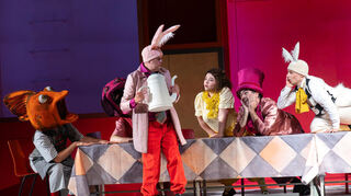 Szene aus der NTM-Oper „Alice im Wunderland“: Die verrückte Teegesellschaft mit (v.l.) Statisterie, Uwe Eikotter (Märzhase), Yaara Attias (Alice), Rafael Helbig-Kostka (der verrückte Hutmacher), Benno Schulz (das weiße Kaninchen).