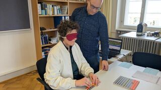 Sylvia M. Felder muss mit verbundenen Augen Aufgaben lösen. Sie wird angeleitet vom stellvertretenden Schulleiter Erich Rüger.